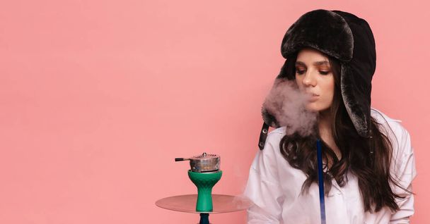 耳フラップとメガネを持つ伝統的なロシアの帽子の魅力的な女性はフックを吸うと喫煙を楽しんでいます。ピンクソリッドカラーの背景. - 写真・画像