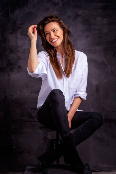 Attractive middle aged woman with toothy smile wearing white shirt and black jeans while sitting against at isolated dark background. Copy space. Studio shot. Full length. - Foto, immagini