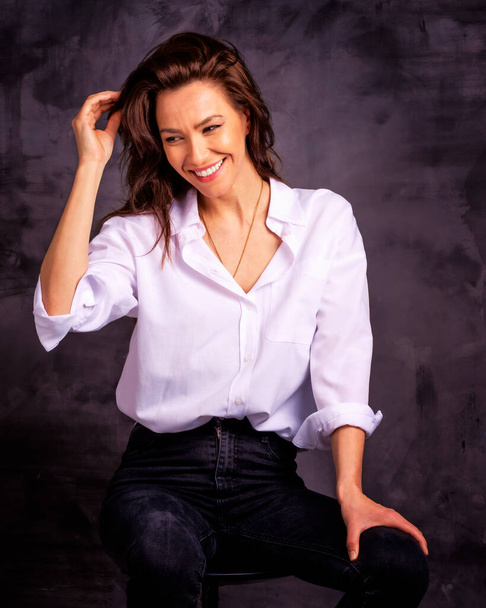 Attractive middle aged woman with toothy smile wearing white shirt and black jeans while sitting against at isolated dark background. Copy space. Studio shot. - Zdjęcie, obraz