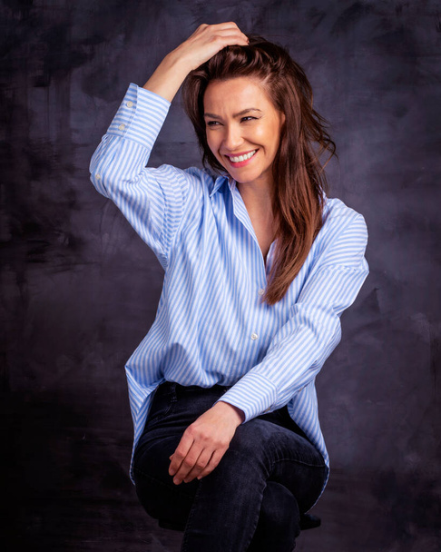 Attractive middle aged woman with toothy smile wearing blue shirt and black jeans while sitting against at isolated dark background. Copy space. Studio shot. - Foto, afbeelding