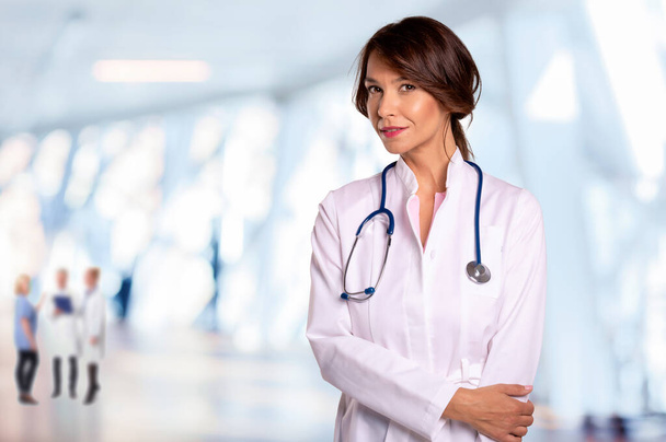 Smiling female doctor wearing lab coat and stethoscope and standing at the hospital corridor.  - Photo, Image