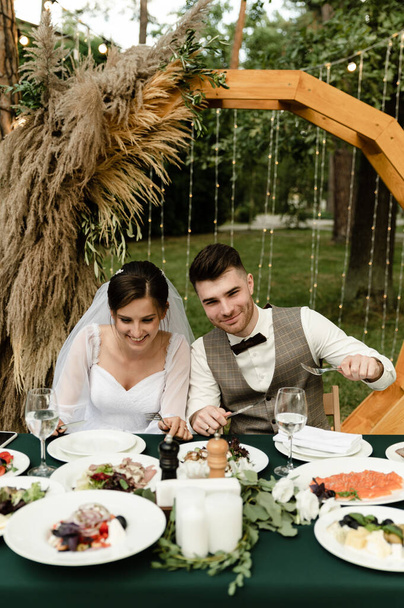 mariés satisfaits sont assis à la table de mariage à l'extérieur. banquet après la cérémonie de mariage. couple de mariage célébrant. traiteur mariage - Photo, image