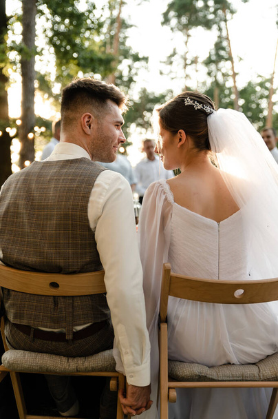 A young groom in a gray suit and a smiling brunette bride are sitting on chairs with their backs to the table, looking at each other and laughing against the background of light bulbs, garlands. Evening portrait. - Photo, image