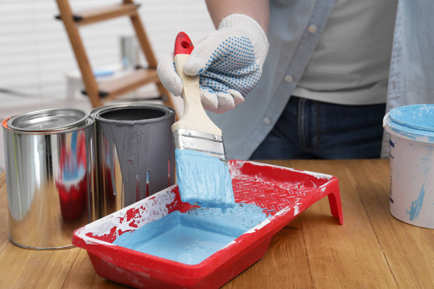 Man taking light blue paint with brush from tray at wooden table indoors, closeup - 写真・画像
