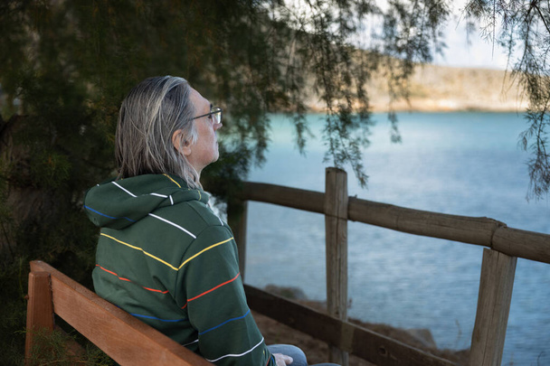 Muž s dlouhými šedými vlasy a brýlemi, sedící sám na lavičce u moře, Řecko, Kréta. Kvalitní fotografie - Fotografie, Obrázek