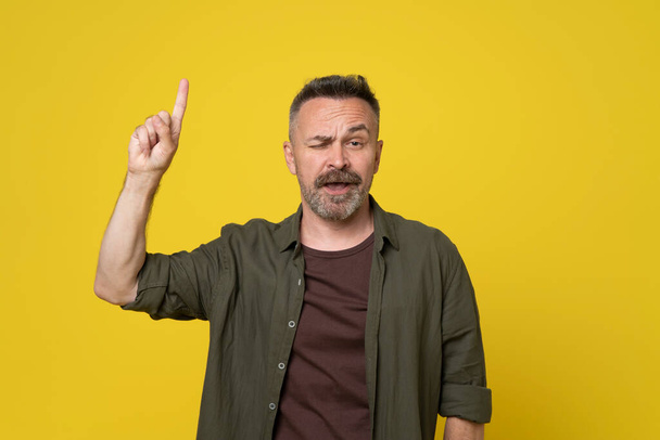 Mature 50s handsome man pointing with finger up above copy space doubtgul grimace on his face with raised eyebrow dressed casual green shirt standing over isolated yellow background - Zdjęcie, obraz