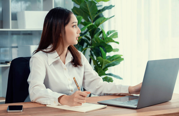 Молодая азиатская восторженная деловая женщина за современным офисным столом использует ноутбук для работы и записи заметок. Усердная и привлекательная офисная леди, работающая над ноутбуком в офисе. - Фото, изображение