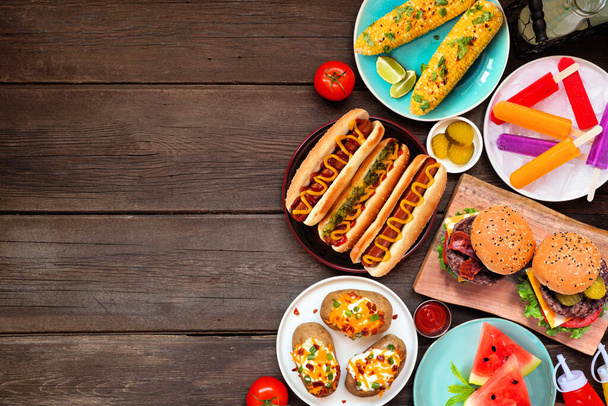 Summer BBQ food side border. Hamburgers, hot dogs, potatoes, corn and cold treats. Overhead view over a dark wood background. Copy space. - 写真・画像