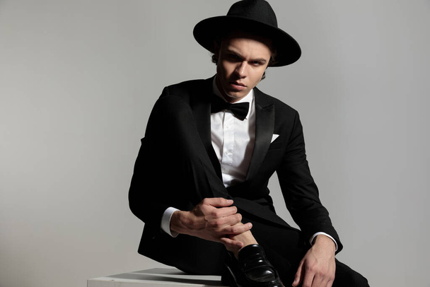 bel homme élégant en tuxeodo noir avec chapeau tenant la cheville et posant devant fond gris en studio - Photo, image
