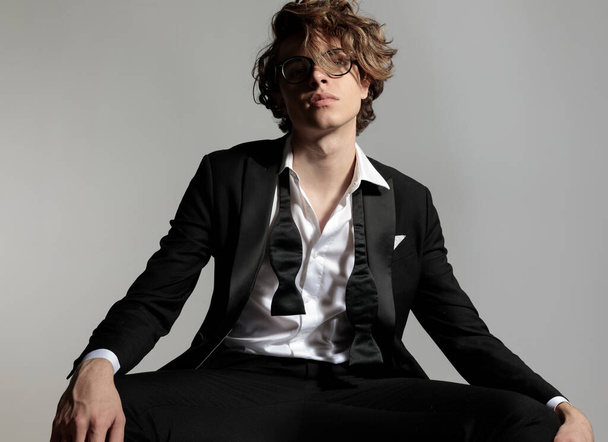 eleganter junger Mann mit Brille trägt schwarzen Smoking und posiert selbstbewusst vor grauem Hintergrund im Studio - Foto, Bild