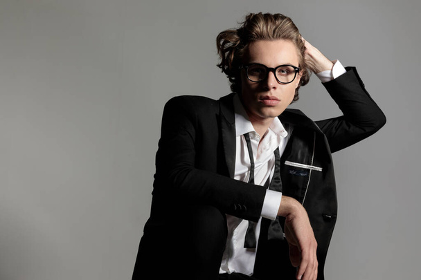elegant young man with glasses adjusting long hair and posing in a cool way in front of grey background - Foto, imagen