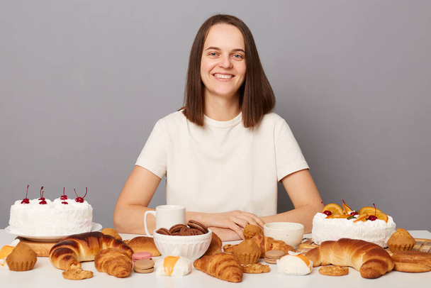 Smiling delighted woman wearing white T-shirt poses at festive table with various desserts, isolated over gray background, looking at camera with positive look. - Foto, Imagem