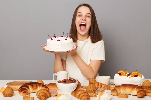Extremely happy satisfied woman wearing white T-shirt isolated over gray background sitting at table holding cake screaming with happiness celebrating with tasty desserts. - Foto, Bild