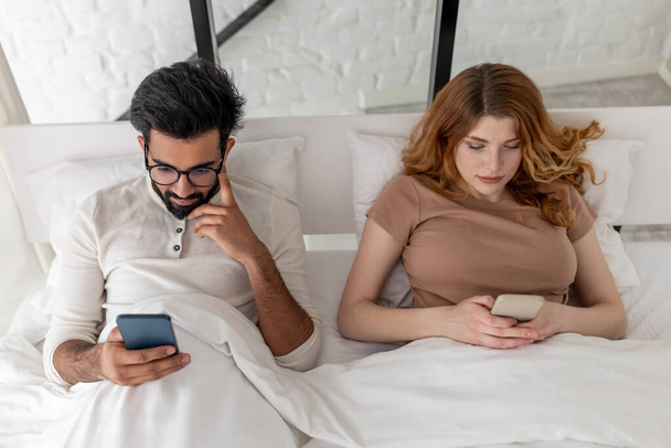 Young mixed race couple with serious expression using smartphone lying on bed. - Valokuva, kuva