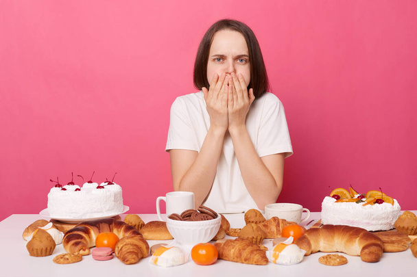 Overeating sick woman wearing white T-shirt sitting at festive table with various desserts, isolated over pink background, covering mouth, being sick, feeling nausea. - Photo, Image