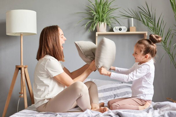 Excited amazed family mother and daughter having fun together playing fighting with pillows laughing sitting on bedroom on bed expressing happiness. - Zdjęcie, obraz