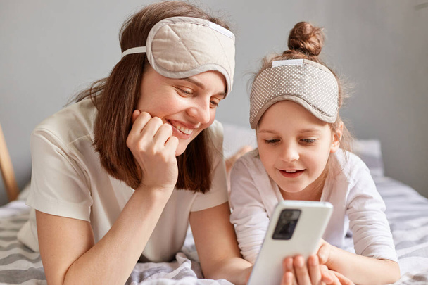 Cheerful woman and her daughter in sleeping mask laying on bed and using mobile phone looking at device screen with smile enjoying checking social networks. - Fotoğraf, Görsel