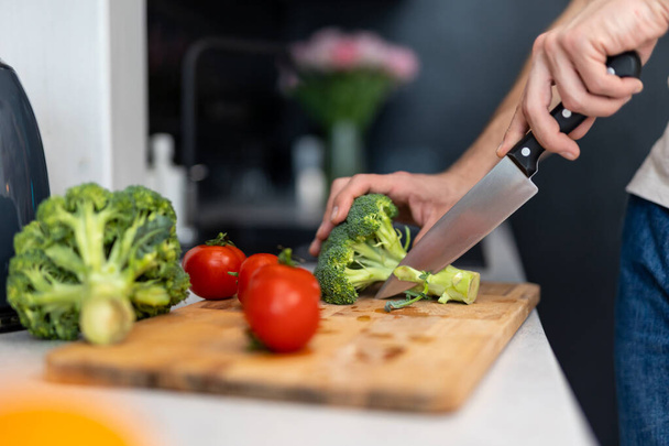 Unknown male hand cuts tomato and brocoli on cutting board with sharp knife in kitchen, making salad - Фото, изображение