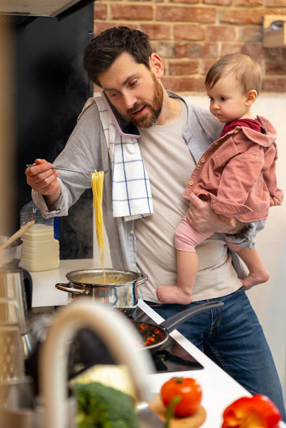 Busy father looking after baby girl making meal and talking on mobile phone, multitasking man at home in kitchenn. - Foto, imagen
