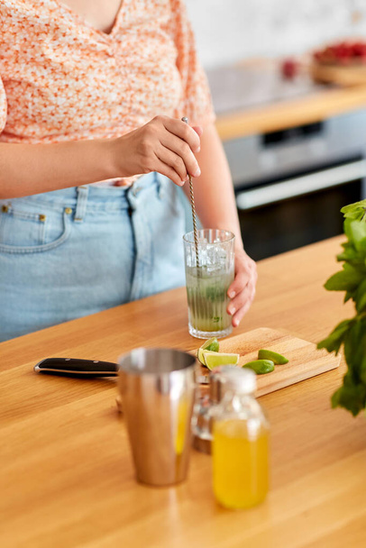 culinary, drinks and people concept - close up of woman with glass and spoon making lime mojito cocktail at home kitchen - 写真・画像