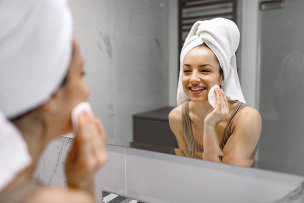 Uśmiechnięta ślicznotka w bieliźnie stojąca w łazience, patrząca w lustro czyszcząc twarz gąbką. Koncepcja pielęgnacji i leczenia urody - Zdjęcie, obraz