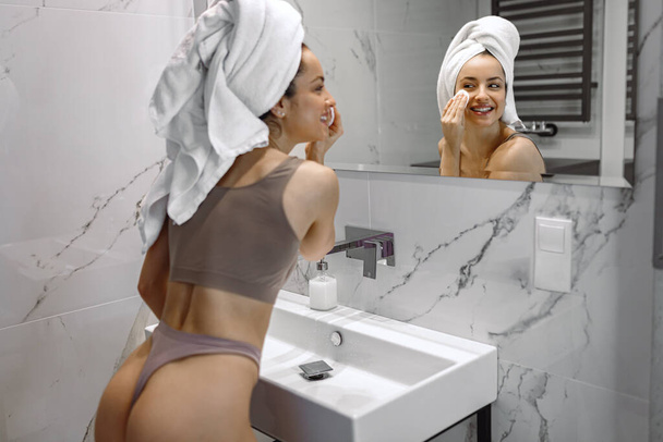 Mutlu genç bayan sabah duştan sonra banyoda dikilirken aynaya bakarken rutin cilt bakımı yapıyor. Güzellik ve Tedavi konsepti - Fotoğraf, Görsel