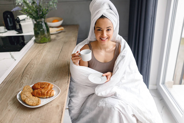 Eine lächelnde Frau, in eine Decke gehüllt, hält eine Tasse Kaffee in der Hand und ruht sich zu Hause auf der Küche aus. Morgenroutine. Lifestyle-Konzept - Foto, Bild