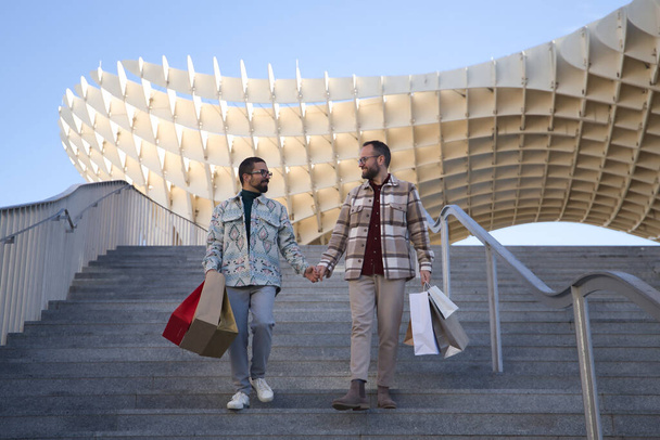 Real marriage of gay couple, holding hands, walking down some stairs and with many shopping bags in the other hand, happy and accomplices. Concept lgtb, lgtbiq+, couples, in love, shopping, sales. - Foto, imagen