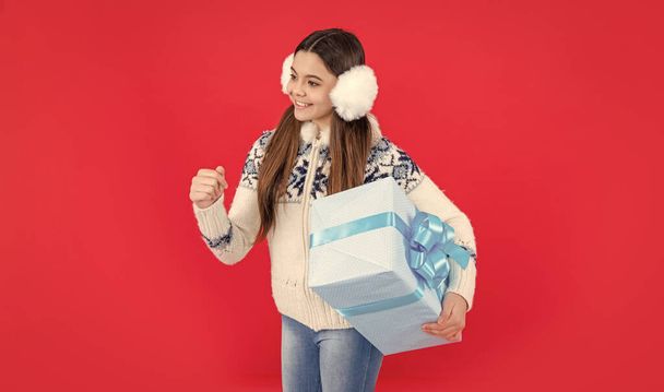 xmas holiday. happy teen girl with xmas present in winter headphones. teen girl hold xmas present isolated on red background. - Photo, image