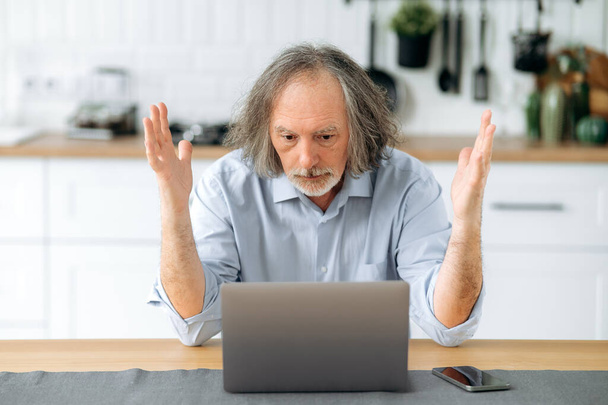 Disappointed caucasian gray haired mature man, pensioner, financial mentor, sit at a table in the kitchen, looks confusedly at a laptop, is perplexed by the news or the result of work, gesturing hands - Foto, imagen