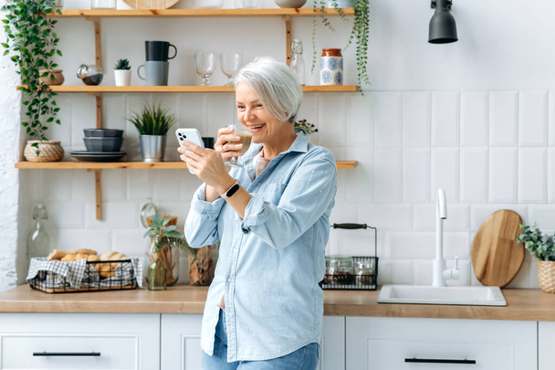 Onnellinen valkoihoinen tyylikäs keski-ikäinen harmaa tukkainen nainen, seisoo kotona keittiössä, pitää älypuhelinta ja lasillista kahvia, viestii sosiaalisissa verkostoissa, lukee uutisia, ottaa selfien, hymyilee - Valokuva, kuva