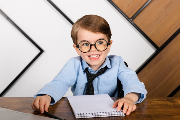 Little boy in round eyeglasses, blue shirt and tie sitting at the desk in the office with a notepad. Smart little kid, Child prodigy. - 写真・画像