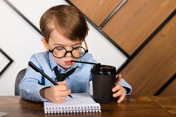 Kleiner Junge mit runder Brille, blauem Hemd und Krawatte sitzt am Schreibtisch im Büro und notiert in einem Notizblock Getränke aus einem Pappbecher. Kluges kleines Kind, Wunderkind. - Foto, Bild