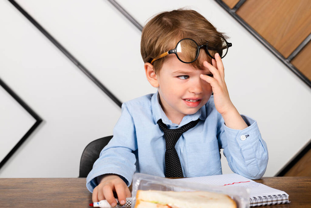 Huilend jongetje in ronde brillen, blauw shirt en stropdas aan het bureau, wrijft in zijn ogen. triest, gestresst kind doet zijn huiswerk. - Foto, afbeelding