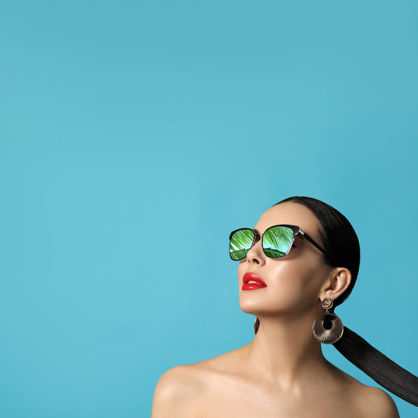 Attractive woman in stylish sunglasses on light blue background, space for text. Palm leaves and sky reflecting in lenses - Foto, Imagen