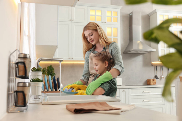 Mother and daughter in protective gloves washing plate above sink in kitchen - Photo, Image