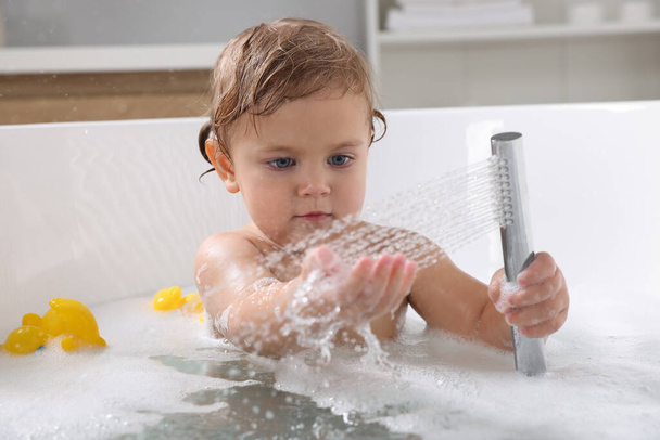 Cute little girl playing with shower head in bathtub at home - Foto, afbeelding
