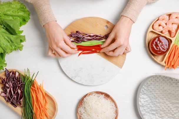 Making delicious spring rolls. Woman wrapping fresh vegetables into rice paper at white wooden table, top view - Foto, Imagem