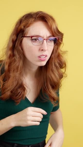 Mujer pelirroja con gafas espiando con curiosidad en estudio con fondo amarillo - Imágenes, Vídeo