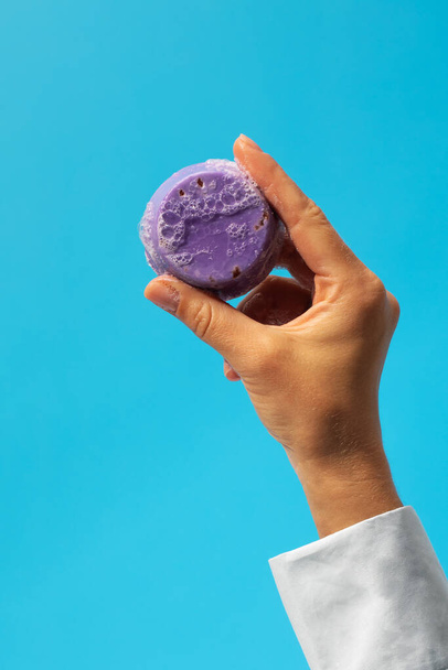 A piece of handmade soap in a womans hand on a blue background. The concept of natural soap, cosmetics. - Fotó, kép