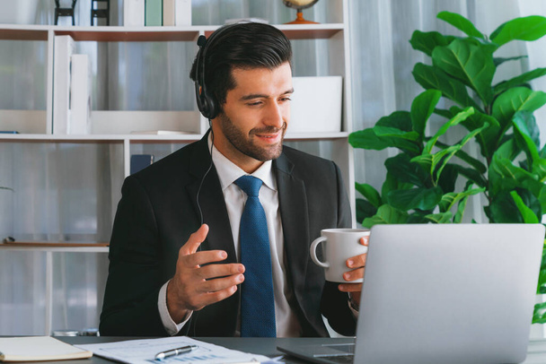 Manager of call center operator office sitting on his desk with his coffee while working on laptop. Office worker wearing headset and black suit working on customer support or telemarketing. fervent - Photo, Image