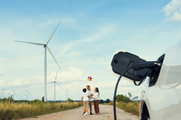 Concept of progressive happy family holding windmill toy and relax at wind farm with electric vehicle. Electric vehicle driven by clean renewable energy from wind turbine generator to charger station. - Foto, immagini