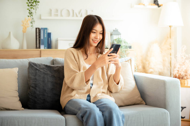 Happy young asian woman relax on comfortable couch at home texting messaging on smartphone, smiling girl use cellphone chatting, browse wireless internet on gadget, shopping online from home - Foto, imagen