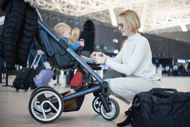 Motherat interacting with her infant baby boy child in stroller while travelling at airport terminal station. Travel with child concept - Photo, Image
