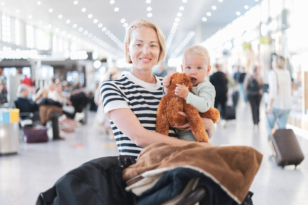 母親は子供と一緒に旅行し、空港ターミナルで飛行機に乗るのを待っている彼の幼児の男の子を抱いています。子供と一緒に旅行するコンセプト - 写真・画像