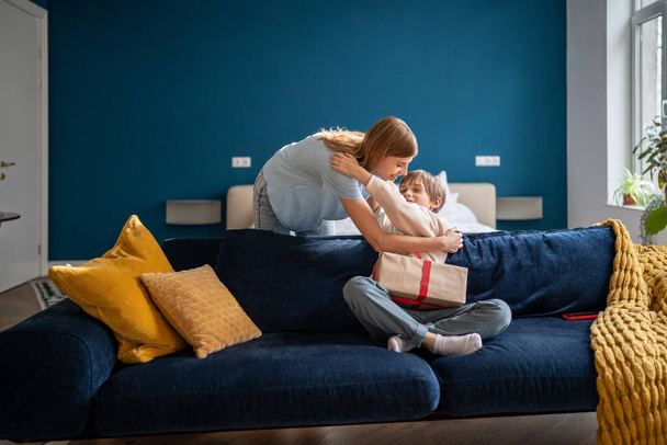 Happy mother giving gift to kid son on birthday, holding present box feeling curious. Excited little son hugging mom on couch at home, going to unpack festive wrap. Motherhood and celebration concept  - Photo, Image