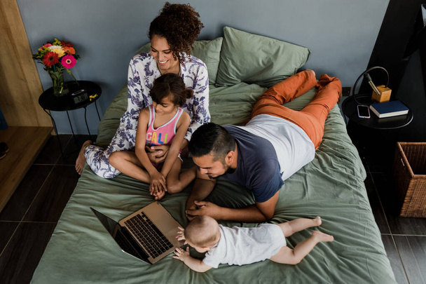 latin family with two children watching movie on laptop or computer together on bed at home in Mexico Latin America, hispanic mother and father - Photo, Image