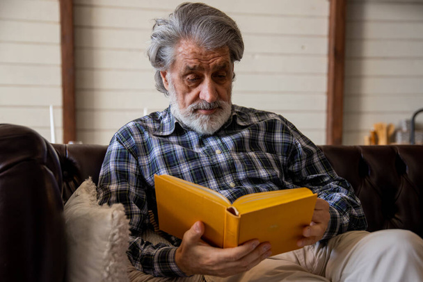 Senior man in checkered shirt reading book while chilling in living room in his house. High quality photo - Foto, afbeelding