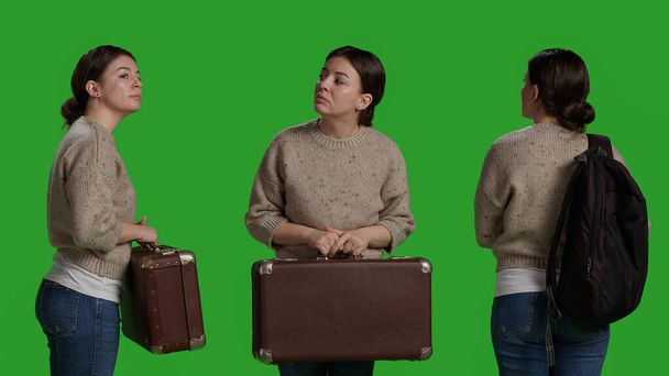 Close up of casual person carrying suitcase and backpack on camera, holding briefcase and luggage to leave on journey. Young confident friendly girl with travel bags, full body green screen. - Photo, Image