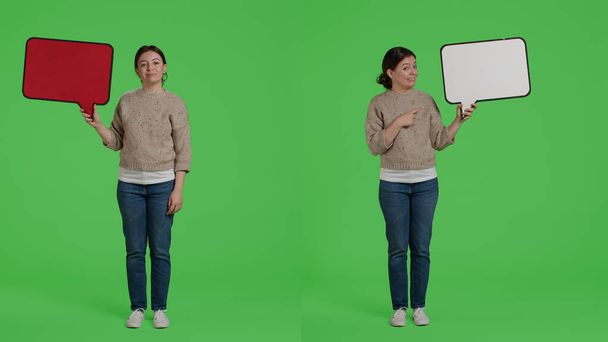Smiling happy girl holding carton speech bubble to create advertisement in studio, showing cardboard icon on camera. Cheerful female model with board standing over greenscreen background. - Photo, image